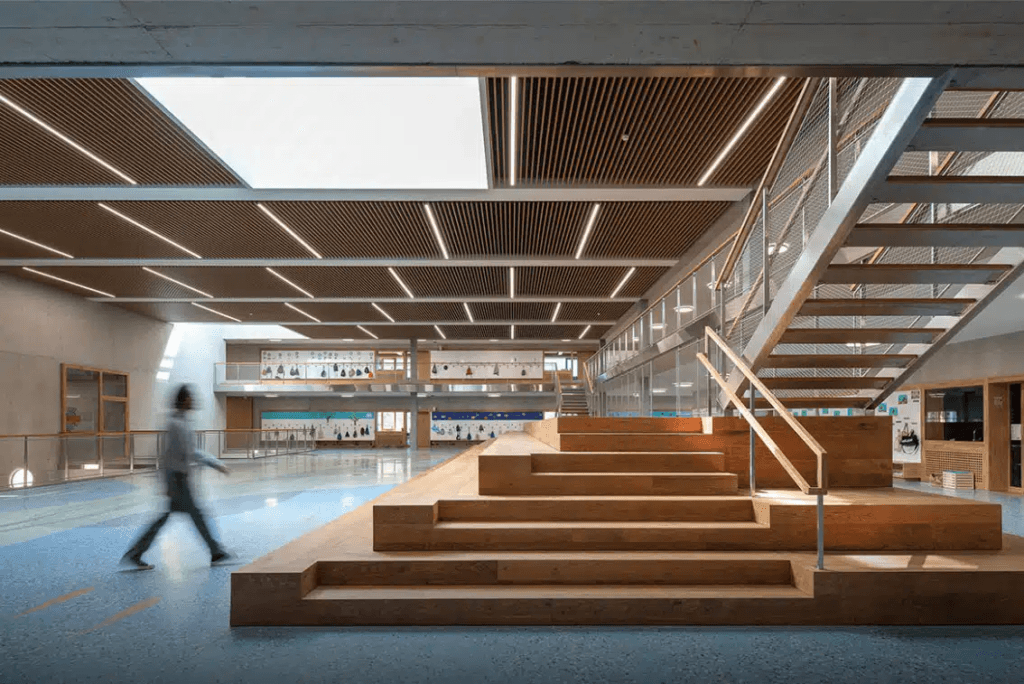 Intérieur moderne avec plafonds suspendus en bois, escalier central en bois avec assises intégrées, et une personne traversant cet espace architectural spacieux et ouvert