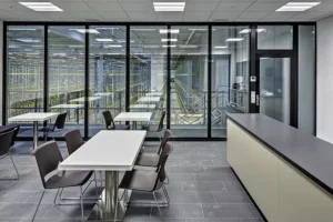 Zone de cafétéria avec des tables et des chaises blanches modernes, dotée d'une paroi vitrée donnant sur un entrepôt high-tech