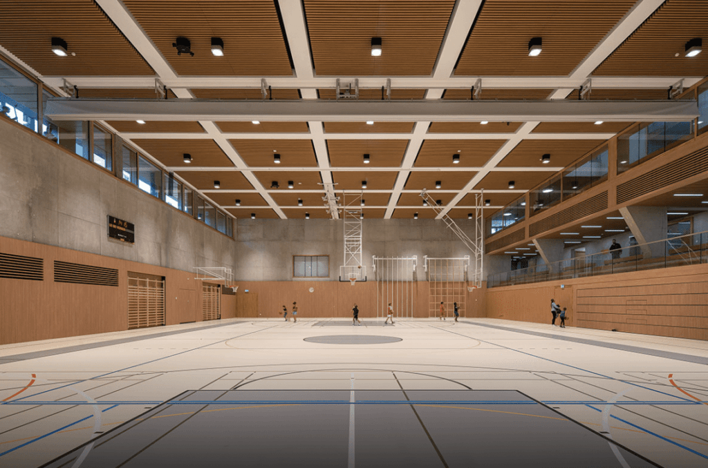 Une grande salle de sport intérieure avec un plafond en bois doté d’un éclairage intégré, un terrain de basketball et des structures d’escalade