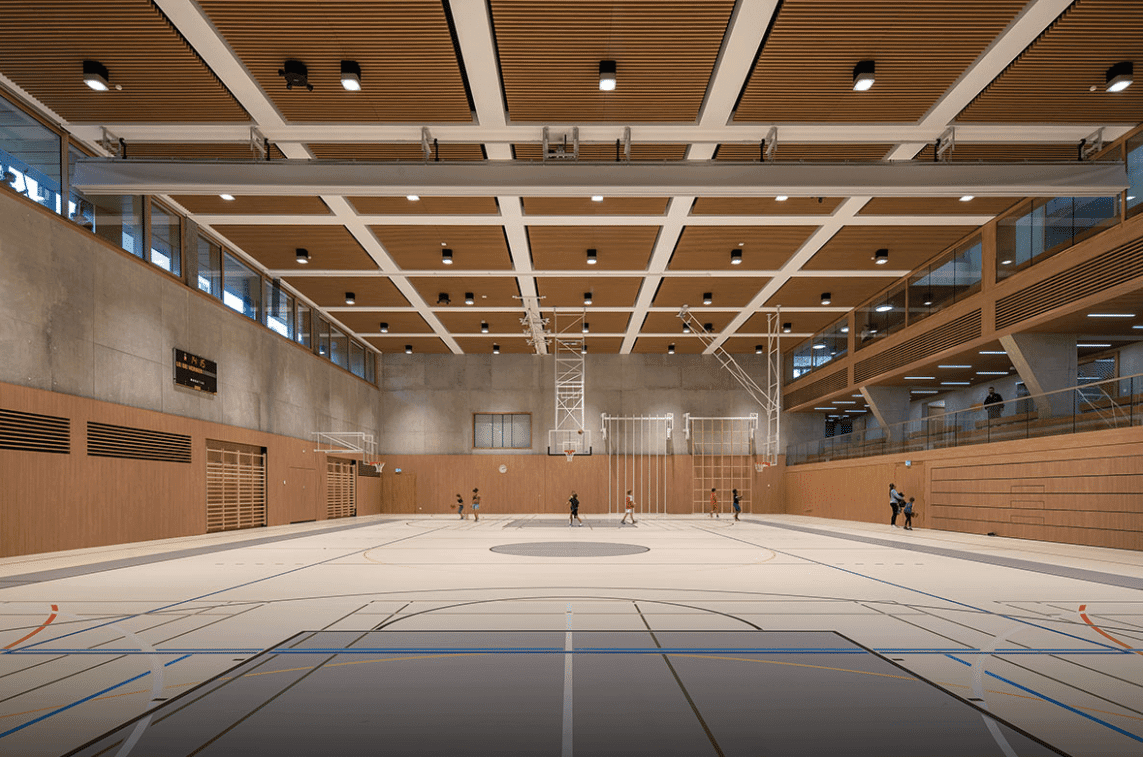 Une grande salle de sport intérieure avec un plafond en bois doté d’un éclairage intégré, un terrain de basketball et des structures d’escalade