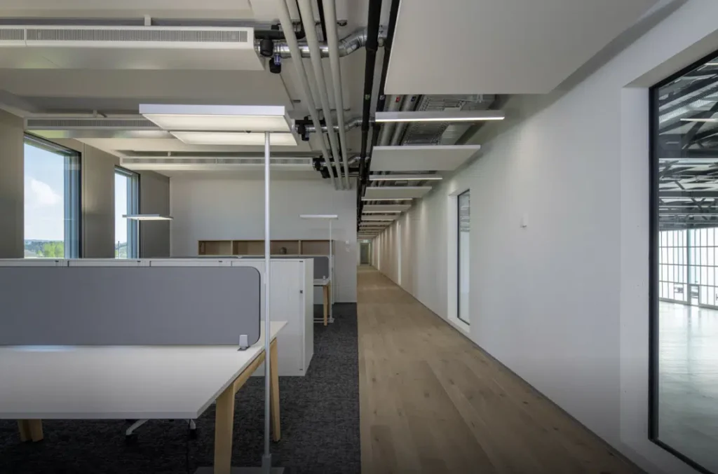Un couloir spacieux de bureau avec des box modernes et des luminaires suspendus au plafond, présentant un design minimaliste avec une lumière naturelle traversant de grandes fenêtres