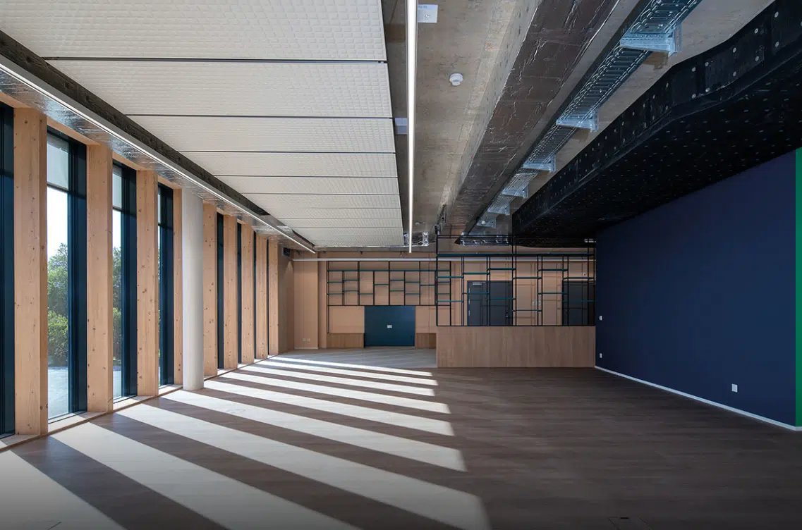 Espace ouvert avec plafond apparent, colonnes en bois et grandes fenêtres, offrant une lumière naturelle et des éléments de design industriel