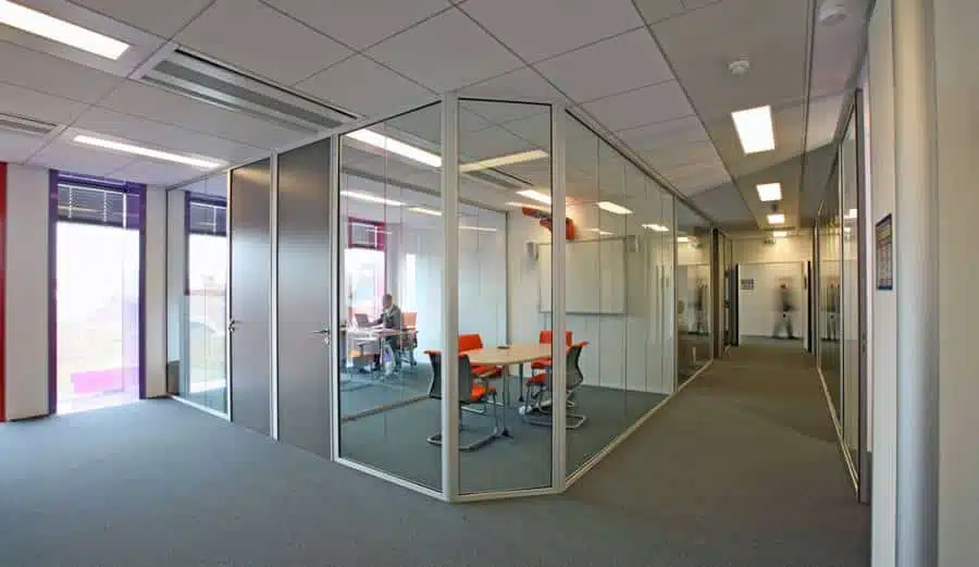 Bureau ouvert avec des cloisons vitrées entourant un espace de réunion, doté de mobilier moderne et d'un éclairage lumineux au plafond