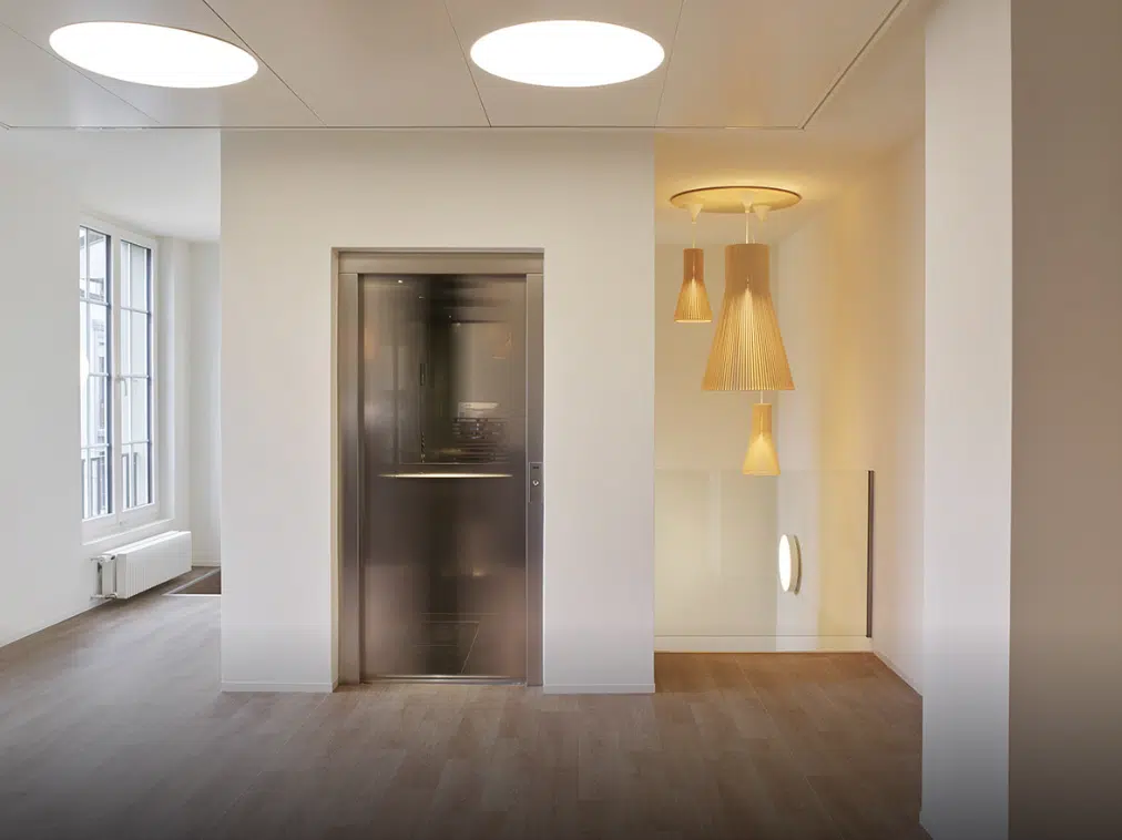 Elevator lobby with wooden flooring, decorative hanging lights, and a clean, minimalist design. French: Hall d'ascenseur avec parquet, lumières suspendues décoratives et design épuré et minimaliste