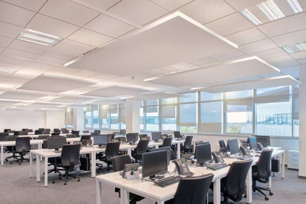 Bureau en open-space avec des panneaux acoustiques suspendus au plafond, de grandes fenêtres et des postes de travail organisés