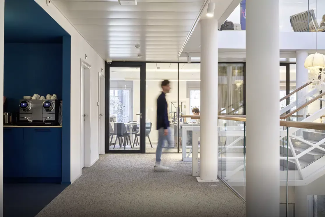 Couloir de bureau lumineux avec une station café, des cloisons en verre et une personne passant, apportant une touche dynamique à l’espace