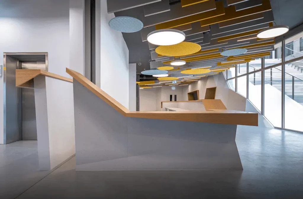 Int&eacute;rieur moderne avec panneaux acoustiques g&eacute;om&eacute;triques au plafond en jaune et bleu, et comptoirs d&rsquo;accueil angulaires en bois